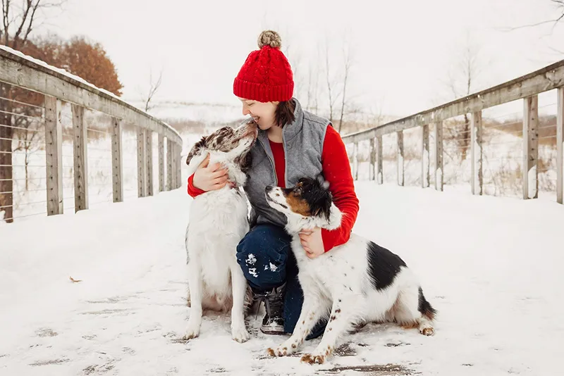 Woman in a red hat hugging two dogs on a snowy boardwalk